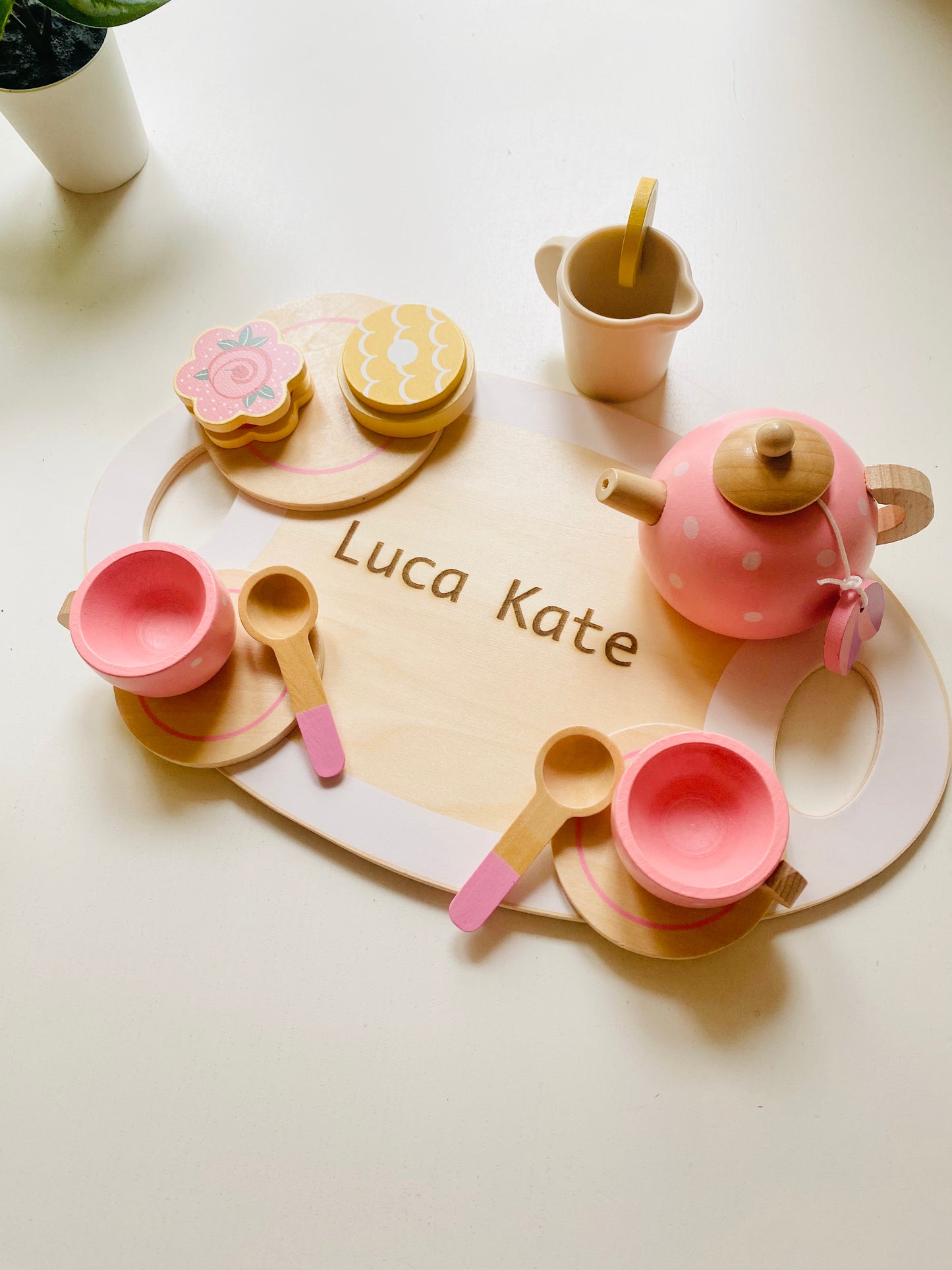 A wooden serving tray with engraving. Two pink wooden cups with saucer and spoons. A wooden pink kettle and a small tray with colourful wooden snacks.A milk pot.