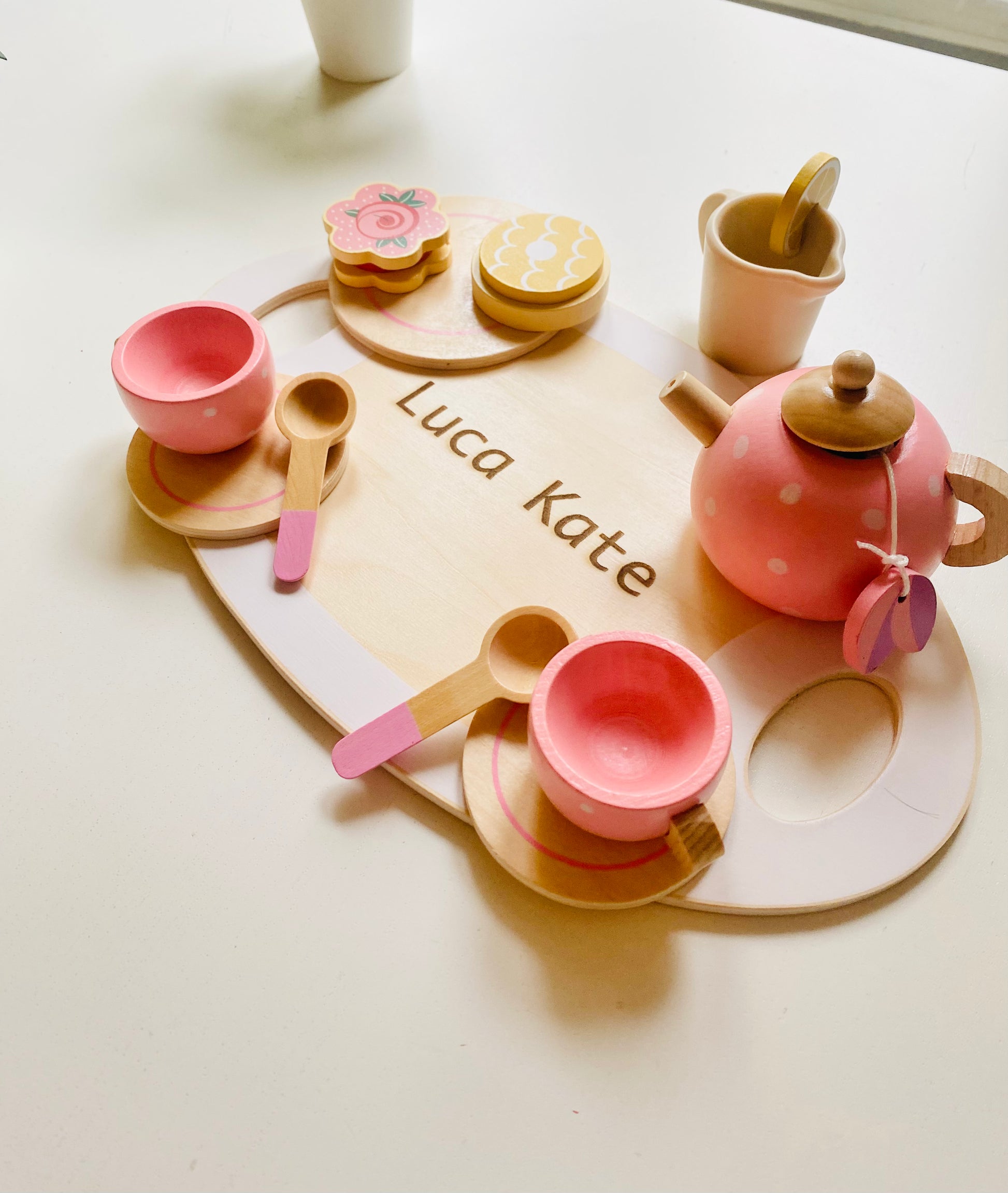 A wooden serving tray with engraving. Two pink wooden cups with saucer and spoons. A wooden pink kettle and a small tray with colourful wooden snacks.A wooden glass lemonade.