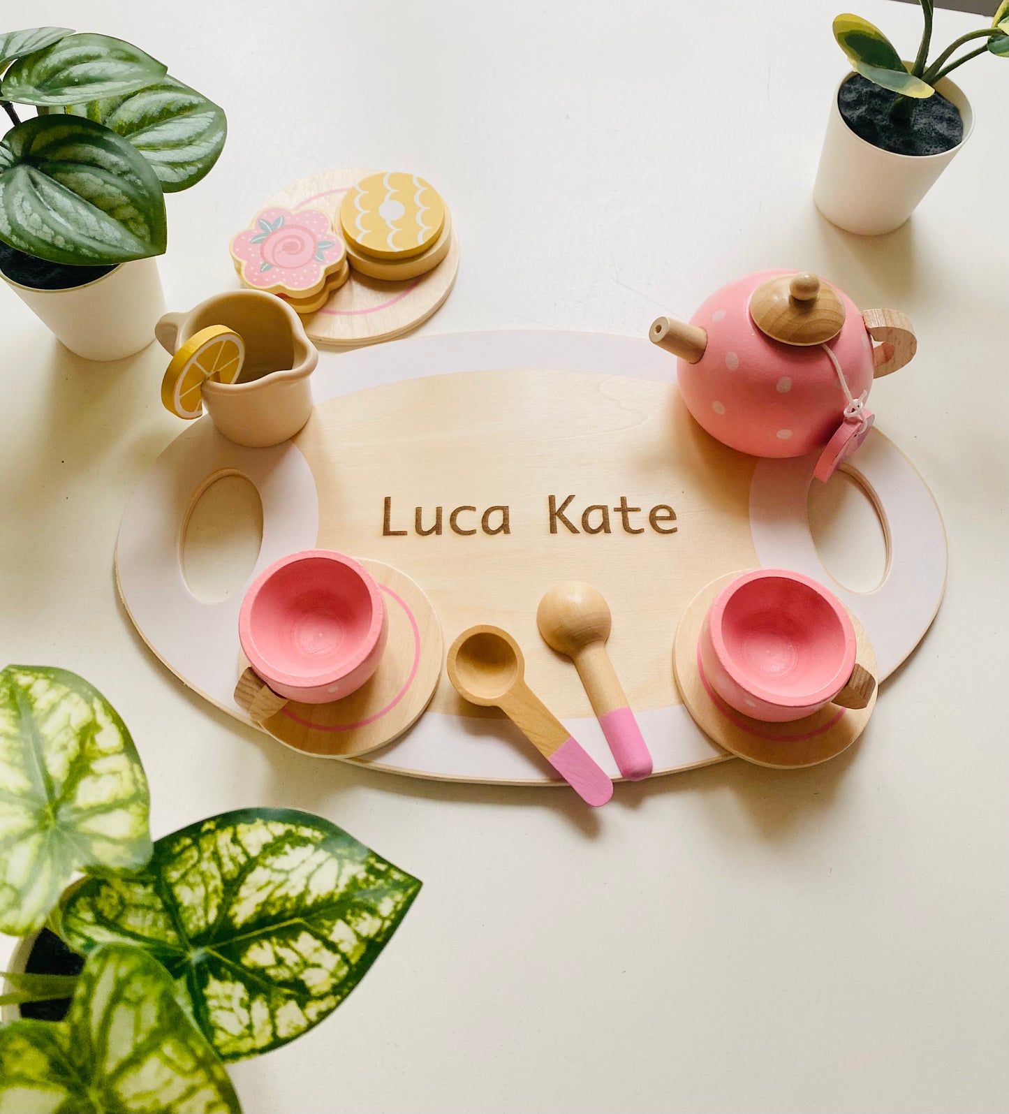 A wooden serving tray with engraving. Two pink wooden cups with saucer and spoons. A wooden pink kettle and a small tray with colourful wooden snacks.A wooden lemonade glass. The picture has 3 plant pot surrounding it.